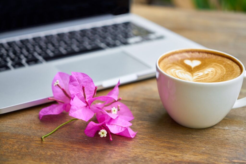 A beautiful latte with heart foam design beside a laptop and bougainvillea on a wooden table.